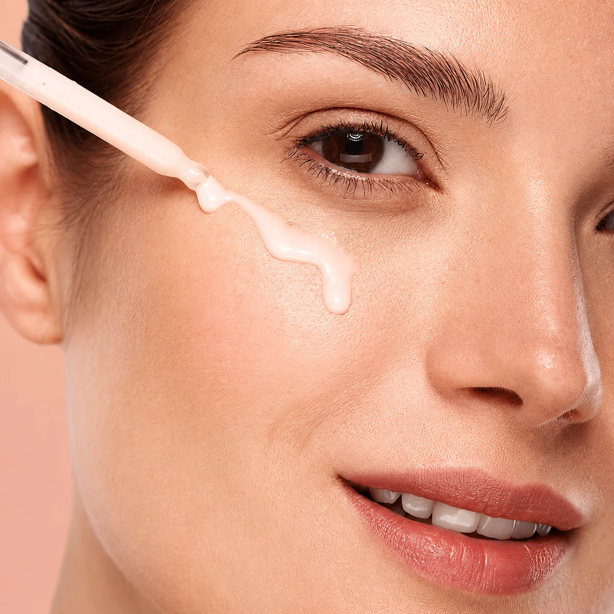 Close-up of a woman's face with liquid serum applied under her eye using a dropper. Glowing skin on a peach background.